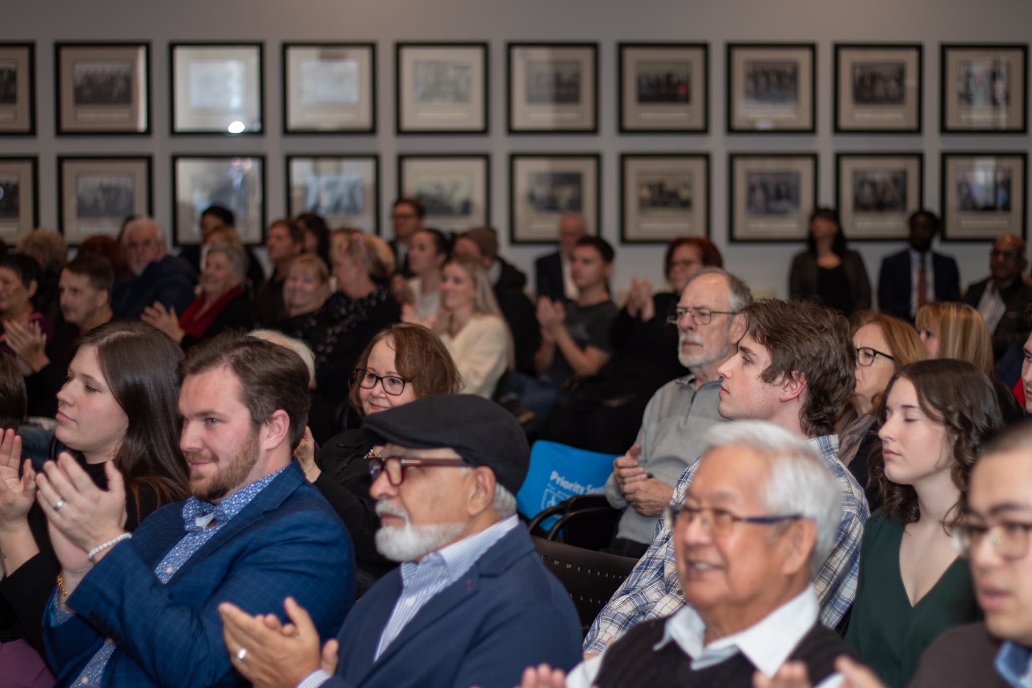 Audience members sitting and applauding at an indoor event, with framed photos displayed on the wall behind them.