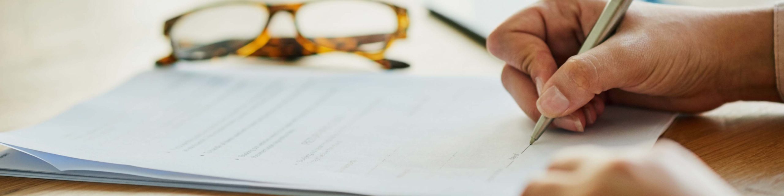 Close-up of a person filling in a form on paper with a pen; spectacles are on the desk in the background.