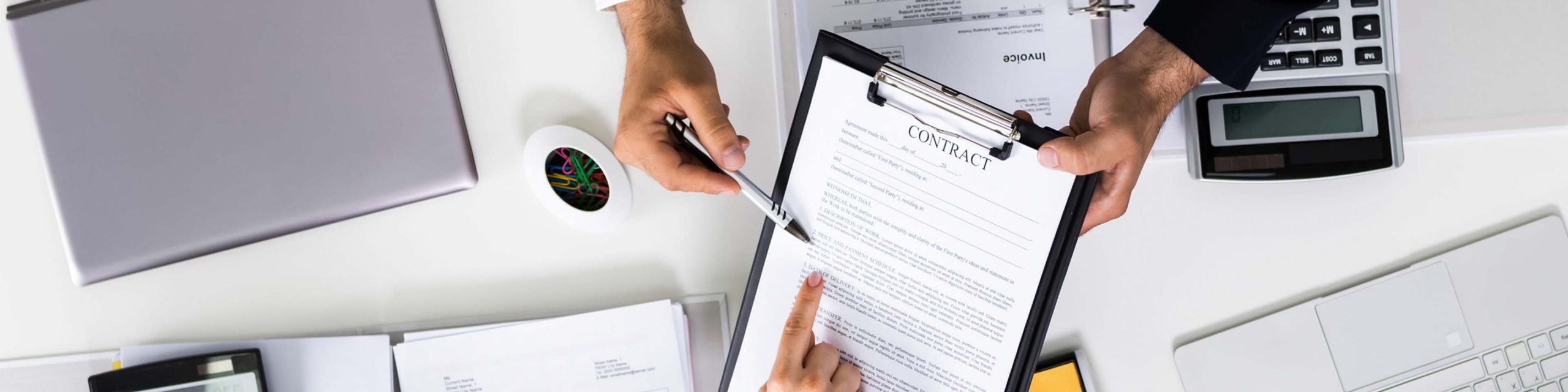 Two people review and discuss a contract on a clipboard at a desk with office supplies, documents, and a calculator.