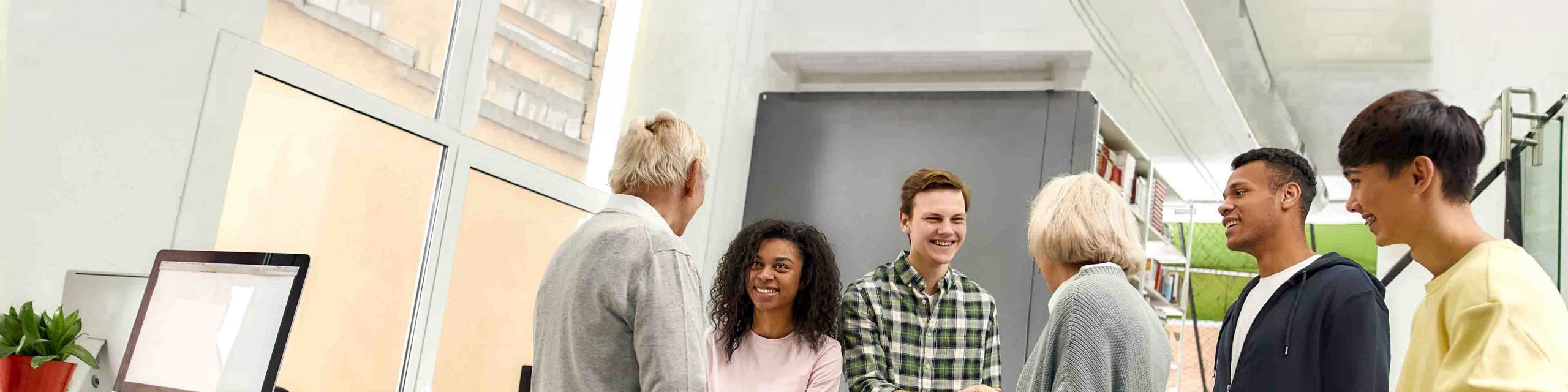 Six people stand and converse in a modern office space near a desk with a computer and green coffee mug.