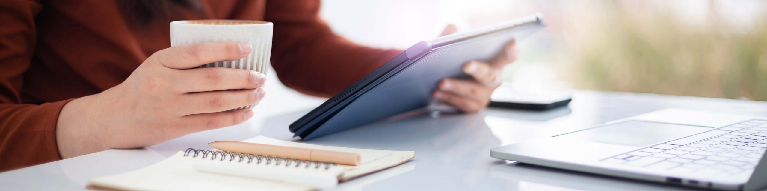 Person holding a cup and a tablet at a desk with a notebook, pen, and laptop.