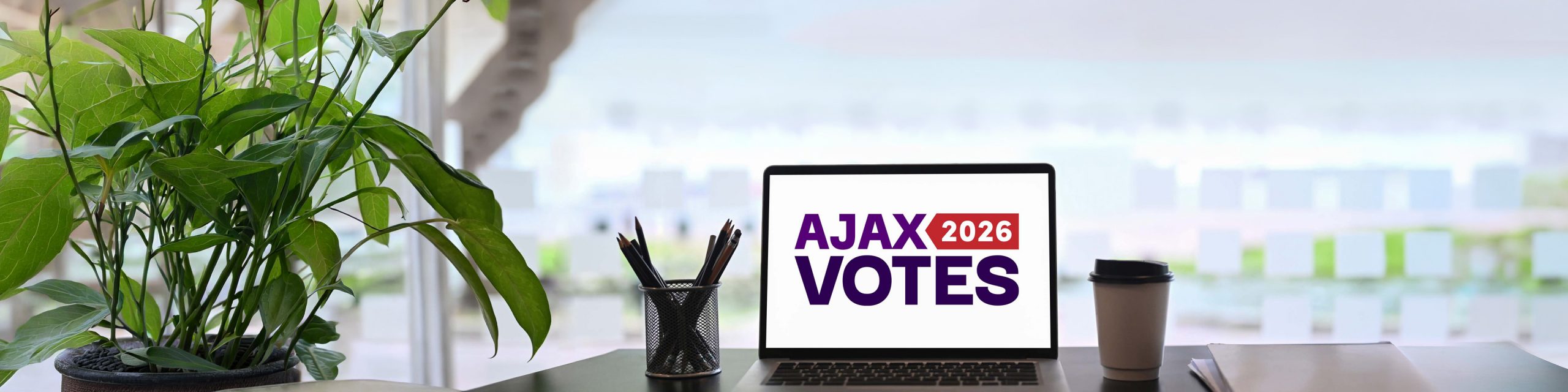 A laptop screen displaying "AJAX VOTES 2026" sits on a desk with pens and a coffee cup nearby.