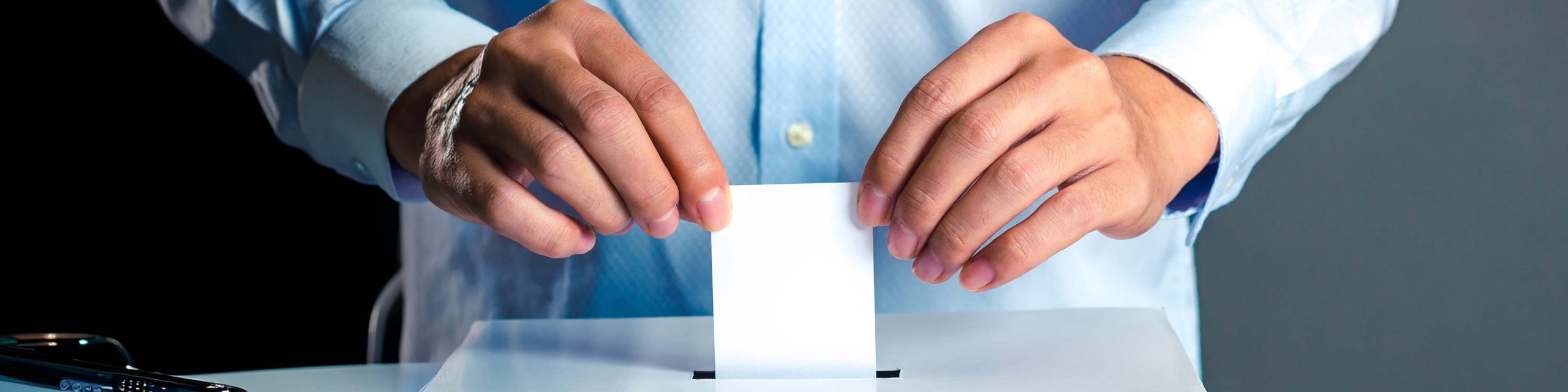 Close-up of a person in a blue shirt inserting a ballot into a white ballot box.